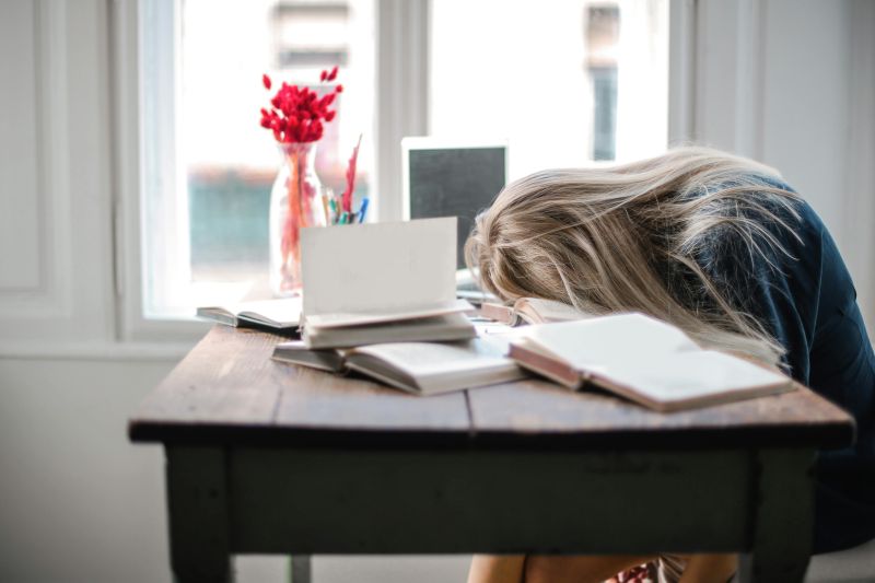 Picture from the office: open dramatic table. Red plants in a vase in the background. A woman sits behind the table with her head propped up on the table.